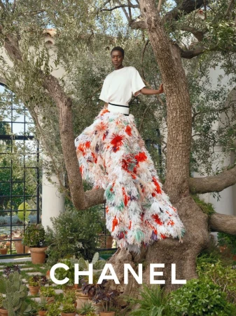Fashion shot of an African woman posing in a floral feather skirt and white top, standing on a tree branch outdoors with Chanel logo.