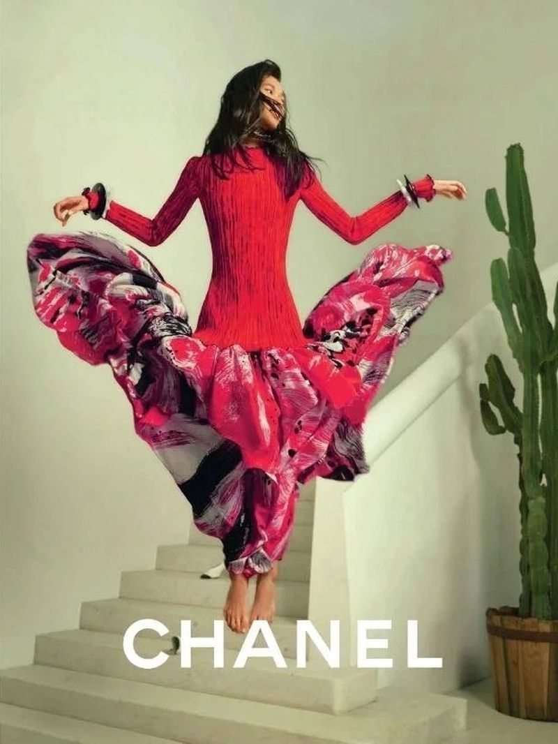 Fashion shot of a woman in a vibrant red dress with a patterned skirt posing on stairs next to a potted cactus, promoting Chanel brand.