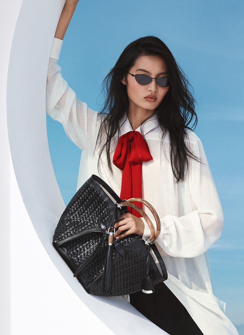 Asian female fashion model poses with black woven handbag, wearing a white blouse, black sunglasses, and a red bow against a blue backdrop.