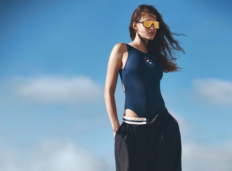 Brunette woman poses in swimsuit and trousers against a blue sky wearing orange sunglasses in a fashion shot in natural lighting.