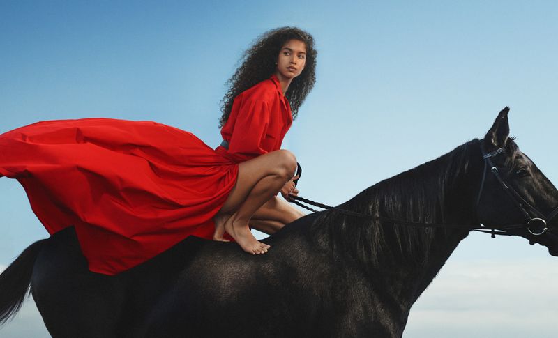 Woman in a red dress riding a black horse in an outdoor setting under a blue sky, equestrian fashion shoot.