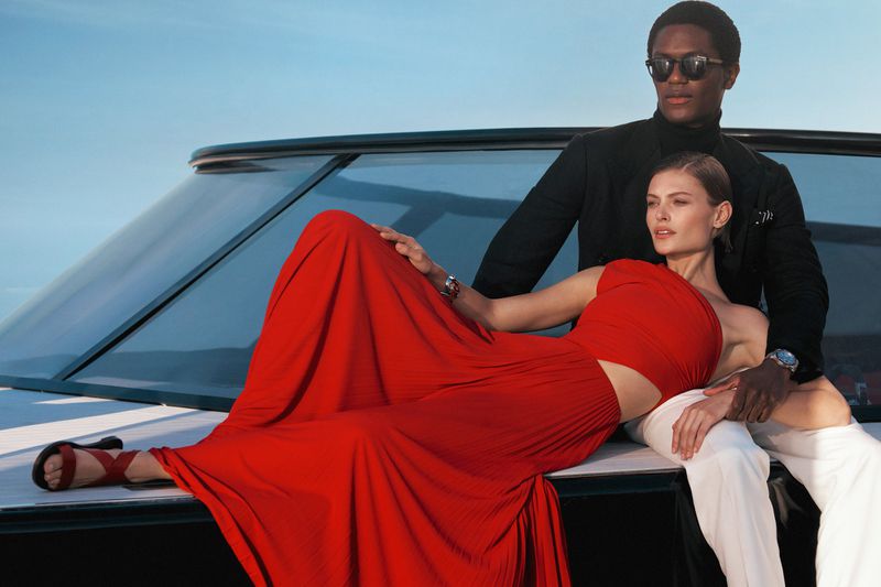 A stylish interracial couple poses on a yacht in the sun. The woman is wearing a flowing red dress and strappy sandals, and the man wears black suit.