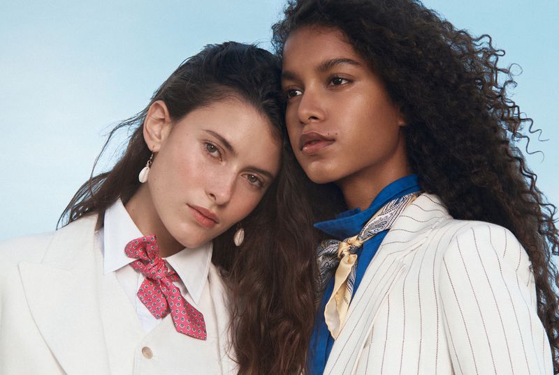 Two adult women pose in tailored white suits with colorful scarves. One woman has dark curly hair, the other has long brown hair.
