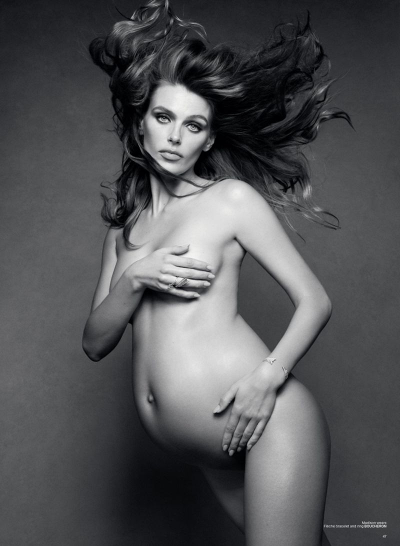 Black and white portrait of pregnant adult woman with wind-blown hair, jewelry, hand poses, in front of gray backdrop.