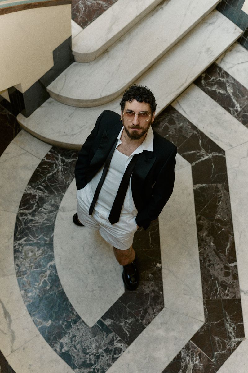 A man in a black blazer, white shorts, and a tie stands on a marble staircase, shot from an eye-level angle, casting a stylish and confident gaze.