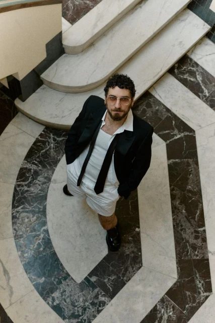 A man in a black blazer, white shorts, and a tie stands on a marble staircase, shot from an eye-level angle, casting a stylish and confident gaze.