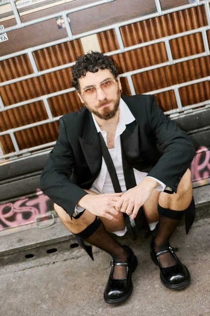 Man in suit jacket, shirt, and shorts poses in Mary Jane shoes, squatting against a graffiti-covered wall with a rolling metal door behind him.