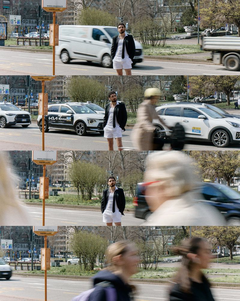 Adult male in white shorts, shirt, and a black jacket standing in the street with cars passing by. Medium shot on sunny day.