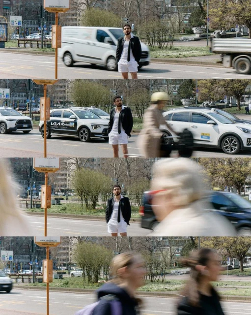 Adult male in white shorts, shirt, and a black jacket standing in the street with cars passing by. Medium shot on sunny day.