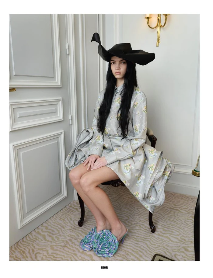 Model seated on a chair wearing a floral patterned dress, dramatic black hat, and patterned mules in an interior room.