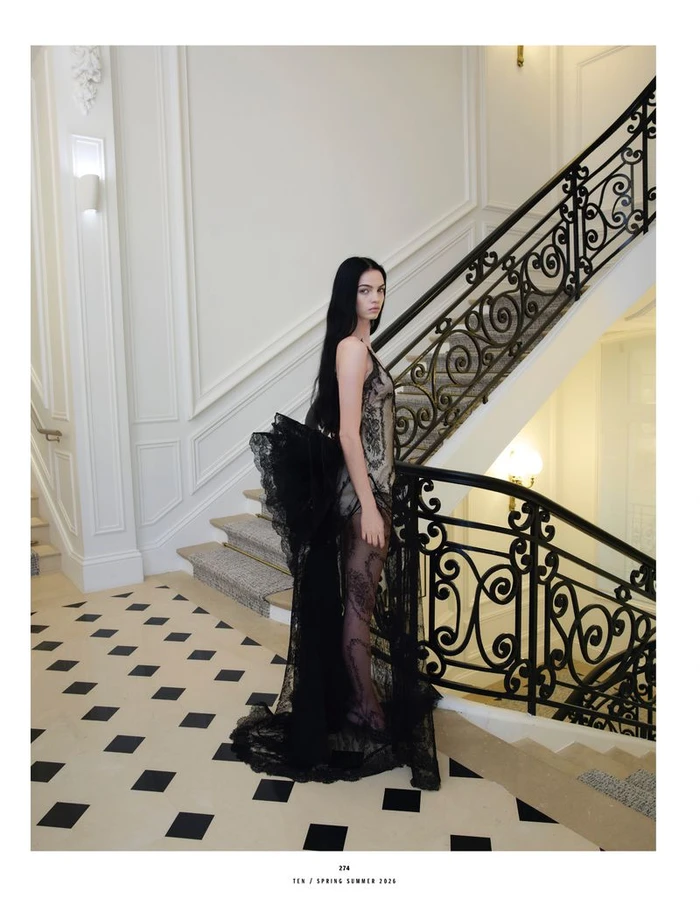 Model standing on a tiled floor in front of an ornate staircase wearing a black lace gown