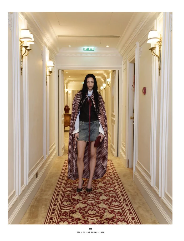 Model standing in a patterned cape and denim skirt in a hotel hallway with ornate wall panels and a red carpet runner.