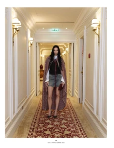 Model standing in a patterned cape and denim skirt in a hotel hallway with ornate wall panels and a red carpet runner.
