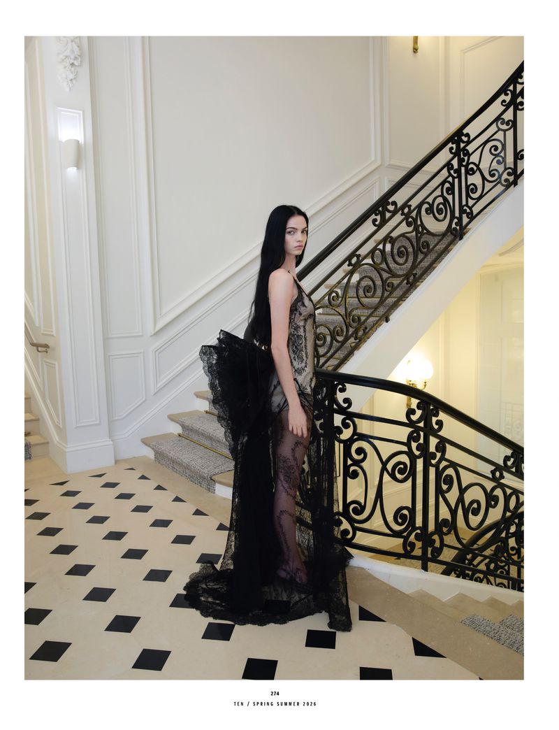 A tall, slender fashion model with long black hair poses on a marble staircase in a sheer black couture gown.