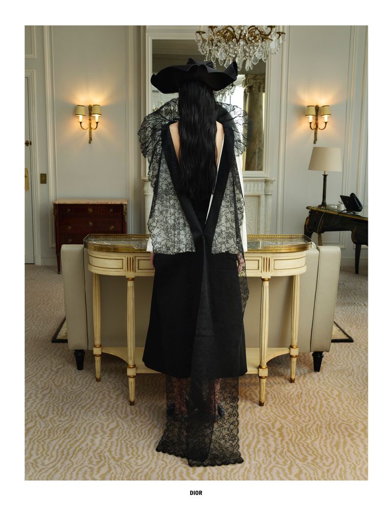 Fashion shot of a woman in a large hat and black gown with lace details, standing in a luxury hotel suite with cream-colored walls.