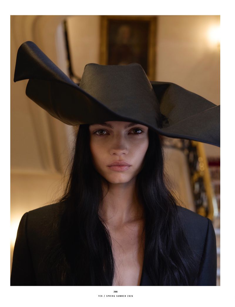 Portrait of a woman wearing a large, black sculptural hat and a black jacket, with a neutral expression, against an indoor setting.