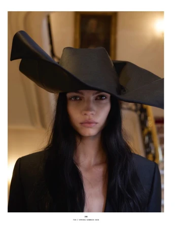 Portrait of a woman wearing a large, black sculptural hat and a black jacket, with a neutral expression, against an indoor setting.