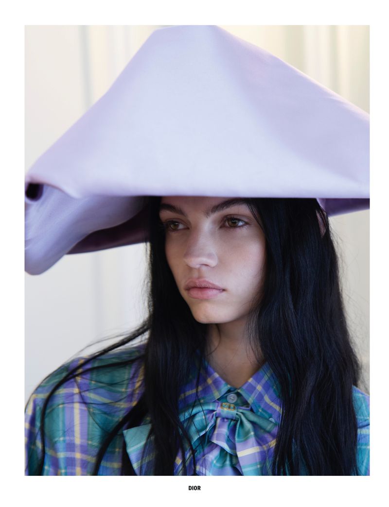Young woman with long dark hair wearing a large, structured lilac hat and a teal and purple plaid shirt with a bow detail.