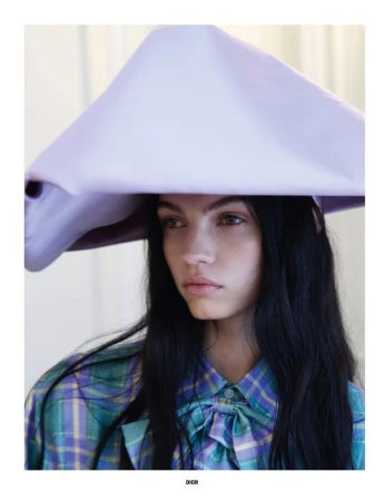 Young woman with long dark hair wearing a large, structured lilac hat and a teal and purple plaid shirt with a bow detail.