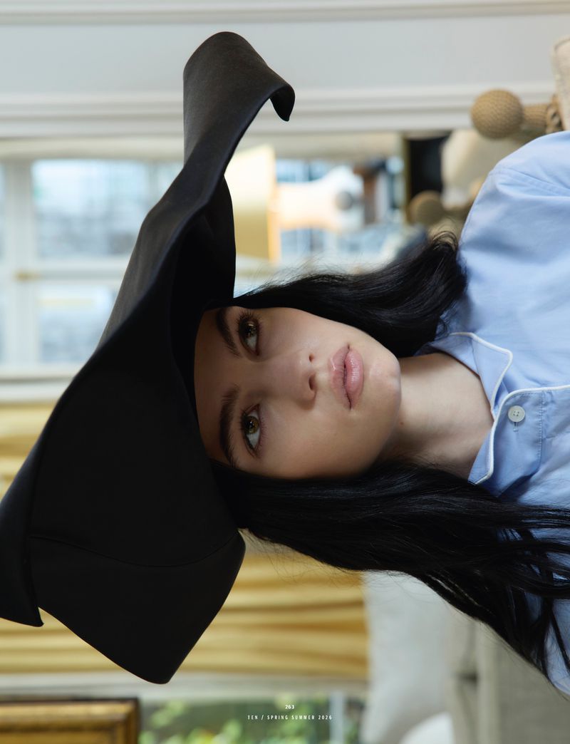 Model wearing a large black sculptural hat and light blue shirt in an indoor setting photographed for 10 Magazine