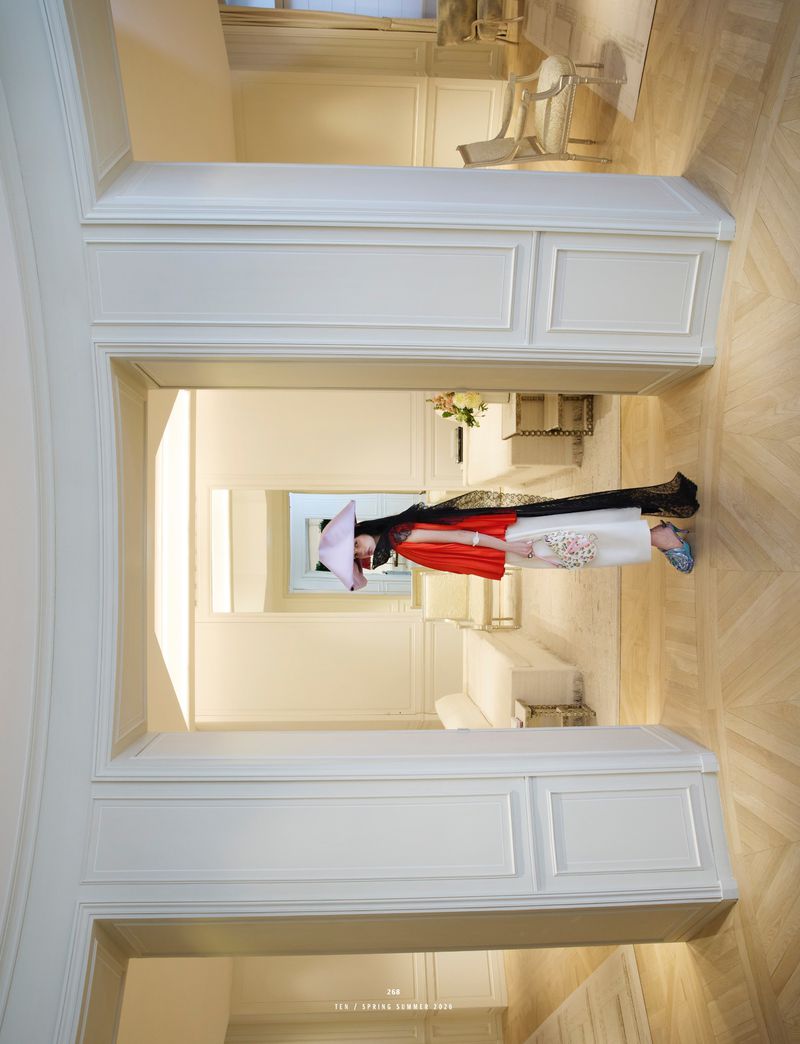 A woman wearing a large hat and fashionable clothing stands in a hallway of a white, modern building with light wood floors.