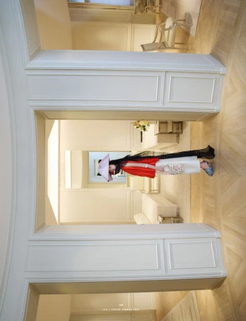 A woman wearing a large hat and fashionable clothing stands in a hallway of a white, modern building with light wood floors.