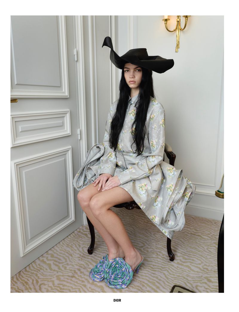 A woman with long black hair poses sitting in a chair wearing a grey floral dress, black hat, and flower-embellished shoes, indoors.