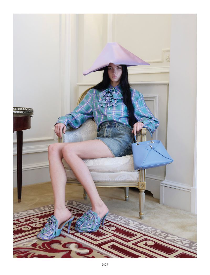 Woman in plaid shirt, denim skirt, cone hat, and floral mules poses indoors with a light-blue purse, sitting on an ornate chair on a patterned rug.
