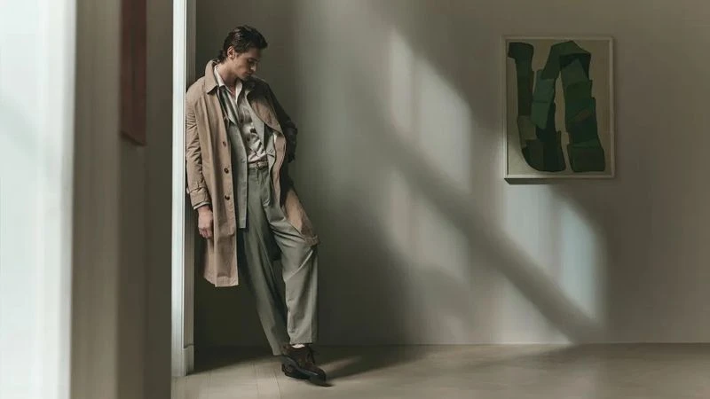 Man in trench coat leaning on wall next to painting. Indoor shot with soft light illuminating the fashion and art.