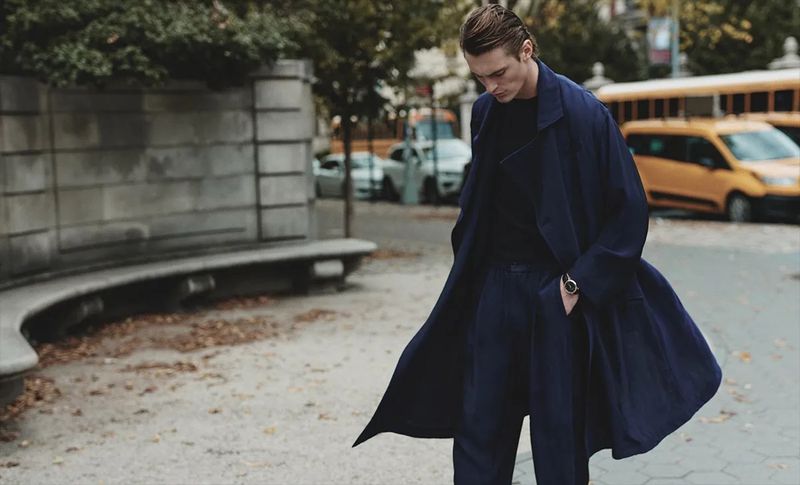 Man in a navy coat walking in an urban setting during daytime. Features include a city park, vehicles, street, and fall leaves.