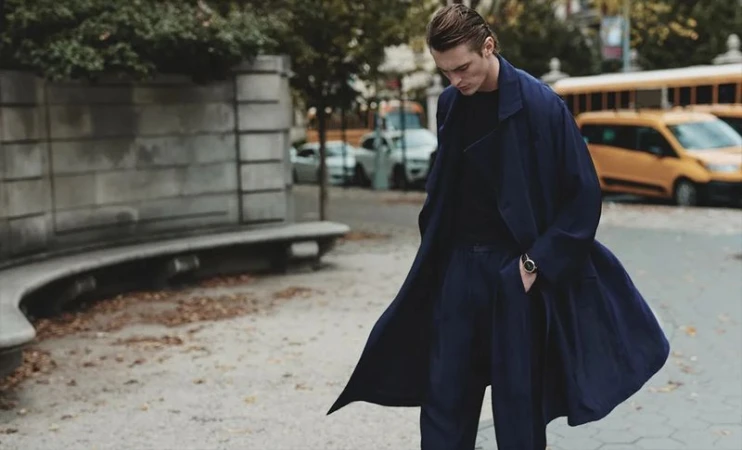 Man in a navy coat walking in an urban setting during daytime. Features include a city park, vehicles, street, and fall leaves.