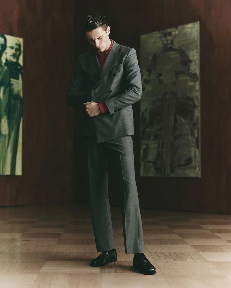 Man in a grey suit and red shirt standing indoors with artwork in the background, looking down in a full shot.
