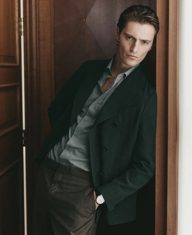 Portrait of a light-skinned adult male in a dark green blazer, light blue collared shirt, and brown pants, leaning against a wooden door.