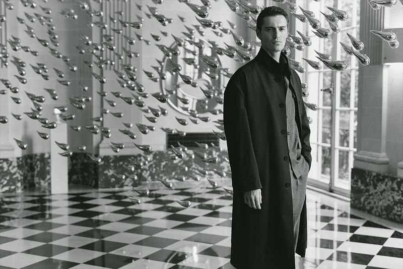 A man in a dark overcoat standing in a grand hall with a black-and-white checkered floor, beneath an installation of silver objects.