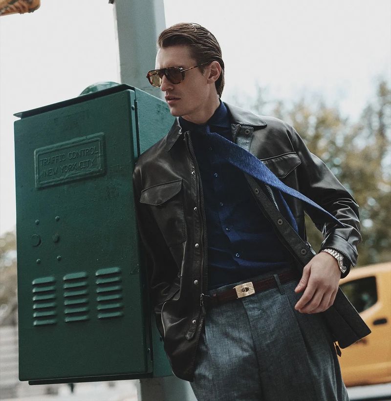 A stylish adult male model in a dark jacket, blue shirt, and grey trousers, poses next to a green traffic control box in urban setting.