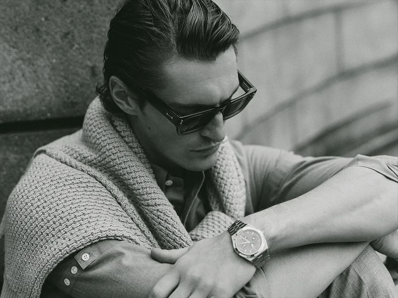 Black and white portrait of a stylish adult male wearing sunglasses and a cardigan, seated outdoors with an intense expression.