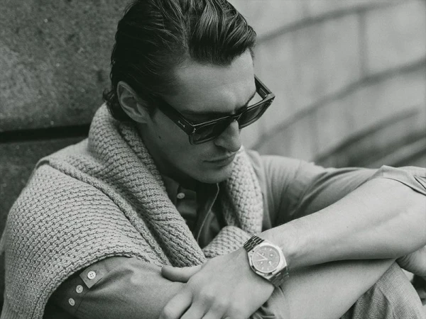 Black and white portrait of a stylish adult male wearing sunglasses and a cardigan, seated outdoors with an intense expression.
