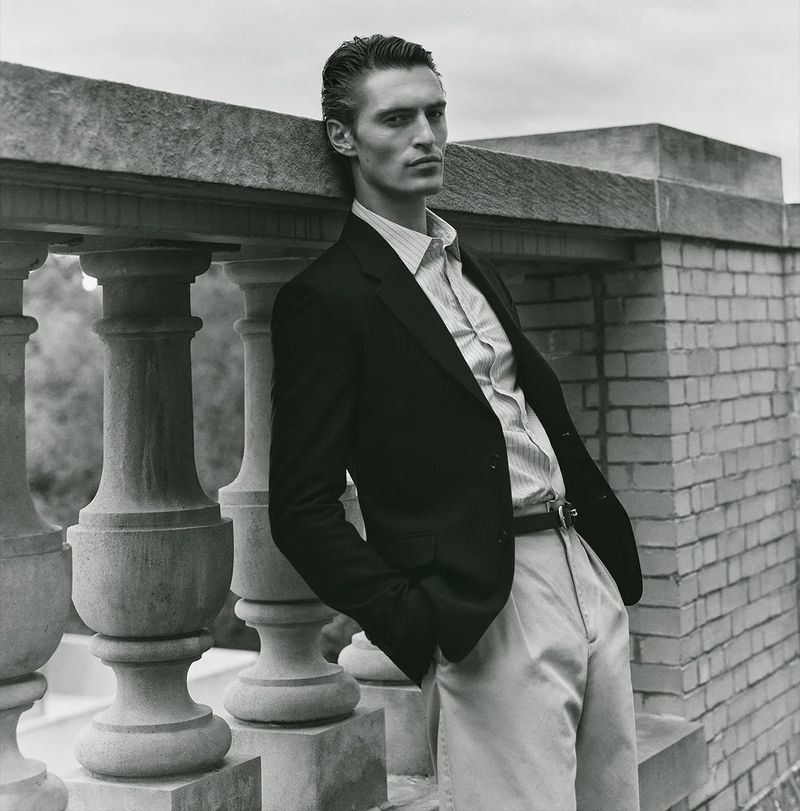 Monochrome portrait of a male model leaning on a stone railing. He wears a dark blazer, striped shirt, and light pants in outdoor setting.