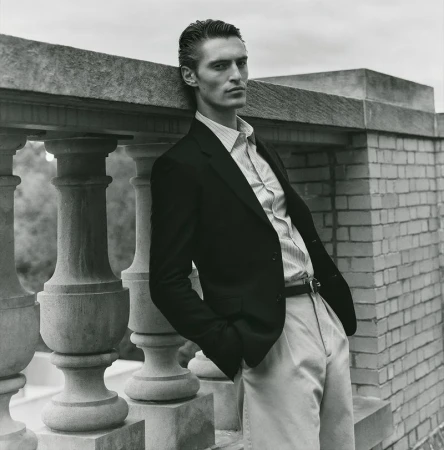 Monochrome portrait of a male model leaning on a stone railing. He wears a dark blazer, striped shirt, and light pants in outdoor setting.