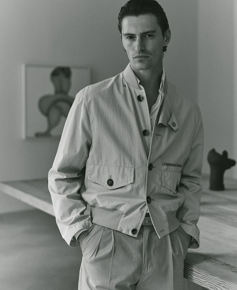A man in a light-colored jacket and pleated pants poses indoors, with art and a wooden table in the background. The image has a neutral tone.