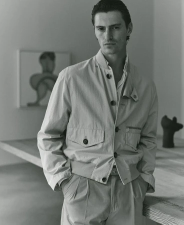 A man in a light-colored jacket and pleated pants poses indoors, with art and a wooden table in the background. The image has a neutral tone.