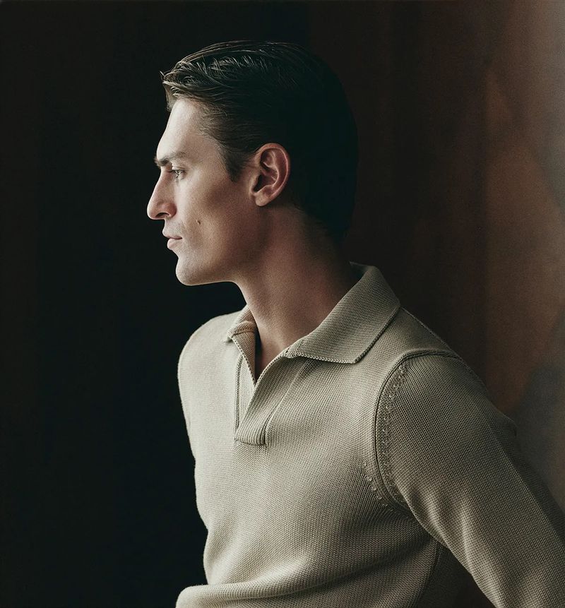 Profile view of a man with dark hair wearing a tan knit polo shirt against a dark background in a studio setting.