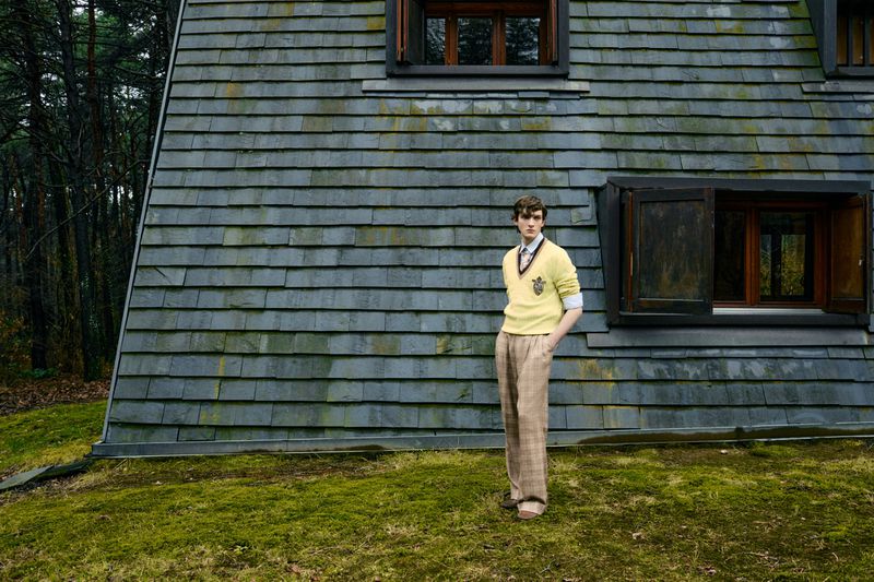 Young man in a yellow sweater and plaid pants stands in front of a mossy, shingled house with dark-wood framed windows and green grass.