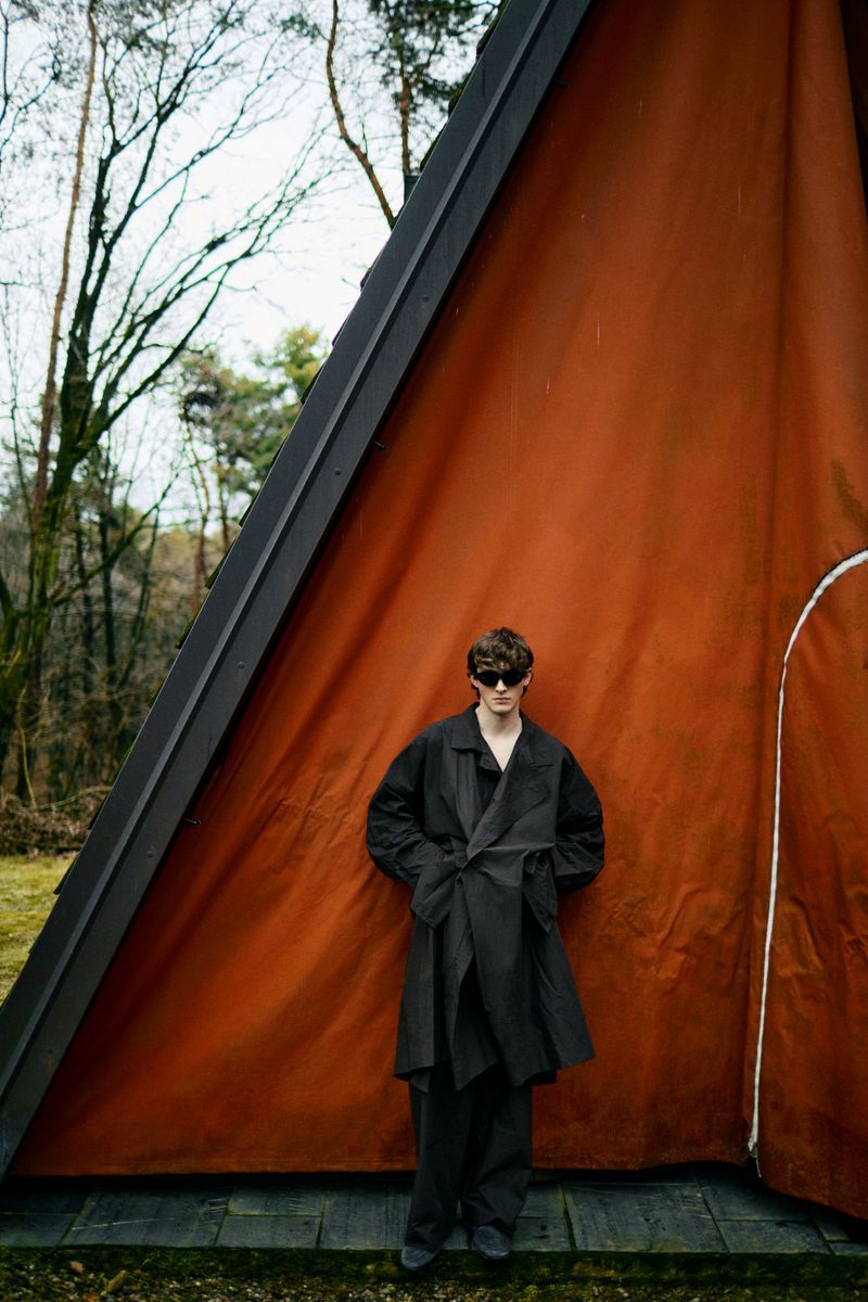Adult male wearing dark glasses and a wraparound coat stands in front of an A-frame cabin on a cloudy day. Outside, fashion shoot.