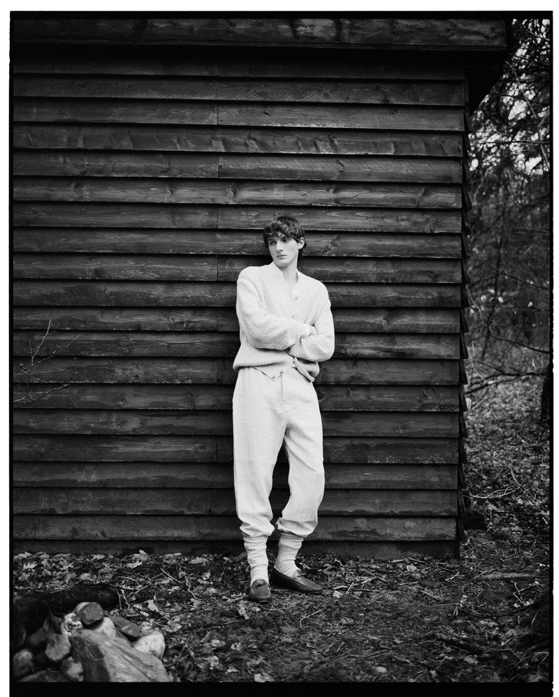 Monochromatic portrait of a young man standing outdoors against a wooden structure, arms crossed, with a forest backdrop and soft, diffused light.