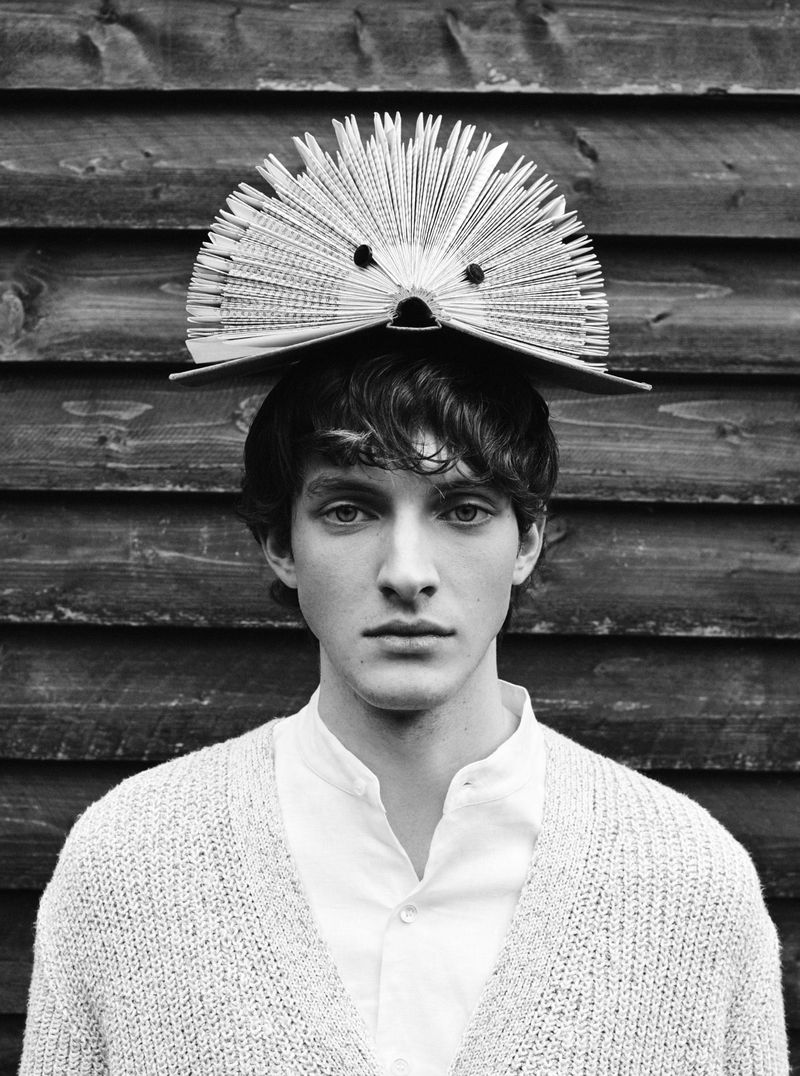 Black-and-white portrait of a young man wearing a cardigan and a book on his head, standing in front of a wood plank wall.
