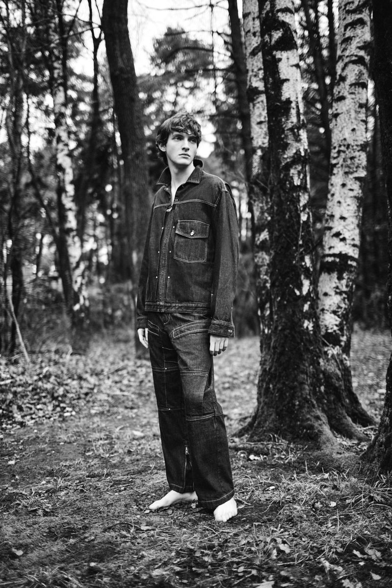 Black and white full shot of a young man in a denim outfit in a forest with birch trees, photographed at eye-level in natural light.