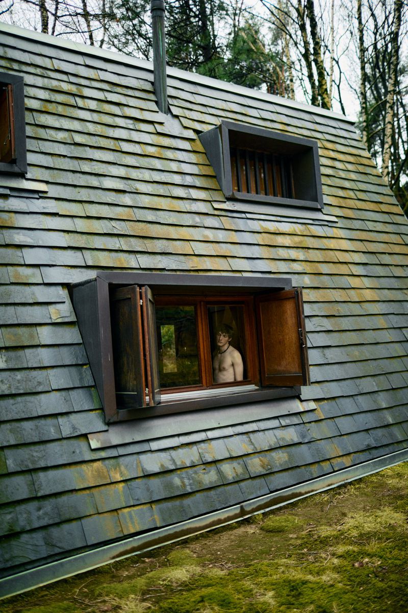 Man gazes from wood-framed window in a unique slate-roofed building, surrounded by trees and mossy ground cover, bathed in soft daylight.