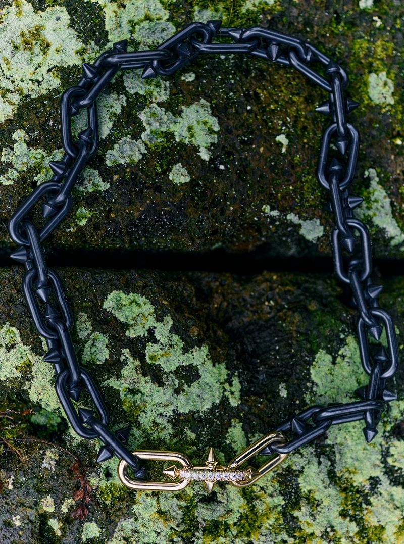 Close-up of a black, spiked chain necklace with a gold and diamond clasp, displayed on a moss-covered rock surface in natural light.