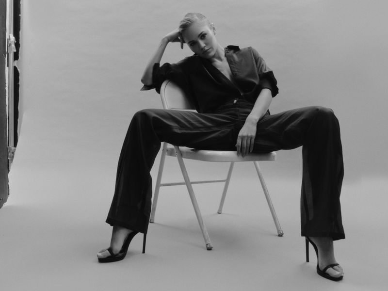 Elegant adult woman in a dark top and trousers poses while seated on a chair, wearing high heels in a studio setting.